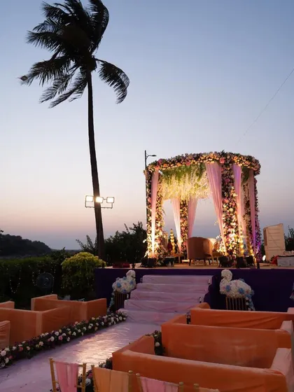 A sunset wedding ceremony with a beautiful floral mandap overlooking the beach. The purple and orange color scheme of the seating and aisle creates a warm and romantic atmosphere.