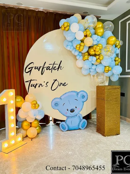 A cute teddy bear theme for Gurfateh's first birthday. This photo backdrop includes a large circular board, a light-up number '1', and a charming blue and gold balloon garland with a teddy bear cutout.