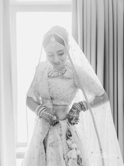 A beautiful black and white getting-ready shot. The backlighting from the window creates a stunning silhouette effect, highlighting her form and the delicate veil.