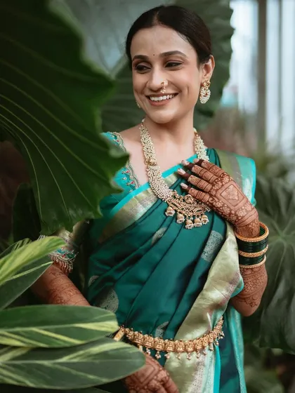 A beautiful portrait of the bride, her smile as radiant as her attire. The styling focuses on traditional elements like the nath and layered gold necklaces, which stand out against the lush green backdrop.