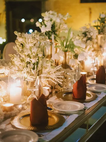 An elegant tablescape for a formal dinner, with gold charger plates, deep red napkins, and a centerpiece of white tuberose, all glowing by candlelight.
