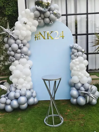 A modern and chic outdoor birthday decoration. This features a custom blue arch backdrop with a personalized hashtag, framed by an elegant silver and white chrome balloon garland.