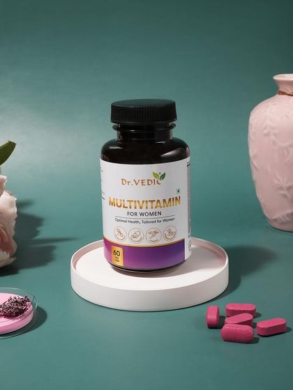 An elegant and clean shot of women's multivitamin capsules. The soft lighting and props like a pink vase and flowers create a gentle and feminine aesthetic.