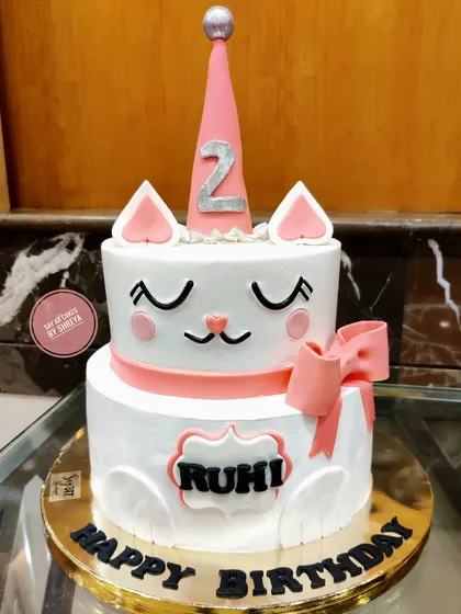 A purr-fectly adorable two-tier cat-themed cake for a second birthday. The design features a cute, sleepy cat face, pink ears, and a party hat, making it a sweet and charming centerpiece.