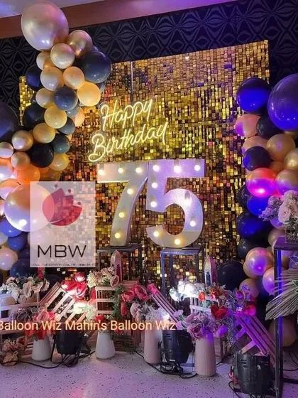 A grand 75th birthday celebration. The stage features a gold shimmer wall, a navy and gold balloon arch, and large, glowing marquee numbers to honor the milestone.
