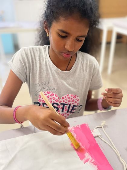 A young girl paints a pink background on her drawstring bag, getting it ready for her main design.