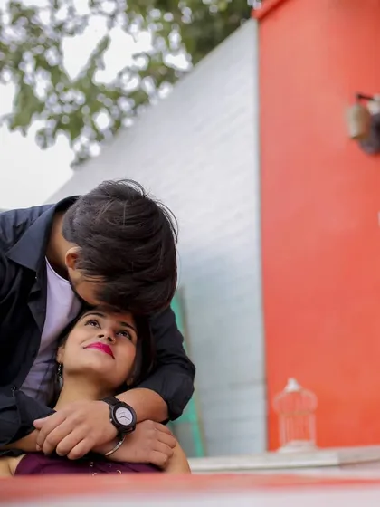 This intimate pre-wedding portrait uses a low angle to create a sweet and adoring perspective. The groom gently kisses the bride's forehead as she looks up at him, capturing a moment of pure affection against a bold, red background.