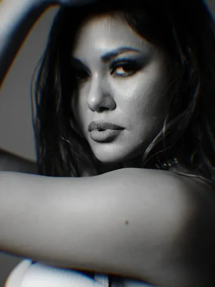 An intense, grainy black and white close-up portrait. The tight framing and dramatic shadows highlight the model's strong features and smoky eye makeup, creating a raw and emotional image.