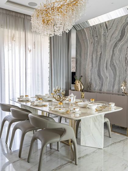 A formal dining room where every detail is considered. The space features a stunning marble table, grey leather chairs, and a dramatic accent wall with book-matched grey marble, all under a beautiful floral-inspired chandelier.