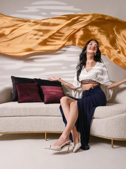 A dynamic and joyful shot for INDY, featuring a model on a modern sofa with a large, flowing piece of golden fabric. This image combines fashion and furniture photography to create a high-energy, aspirational lifestyle scene.