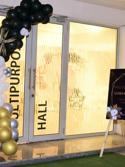 The entrance to a multipurpose hall decorated for a party, with a black, gold, and silver balloon garland and a welcome sign for the event.