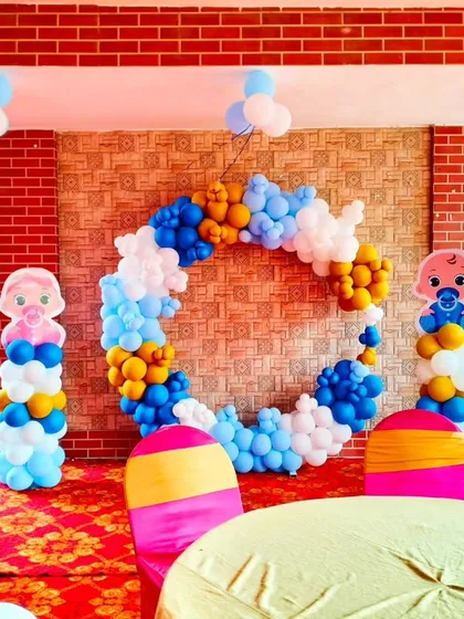 A bright and cheerful setup for a Kuan Pujan ceremony. The decoration includes a circular balloon ring in blue, white, and yellow, with cute baby-themed pillars on each side.