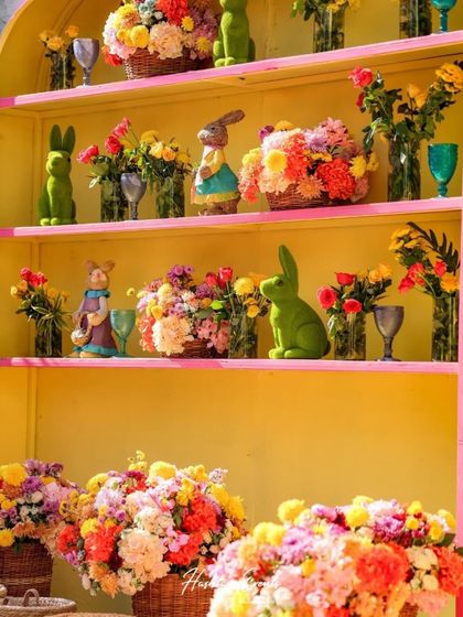 A bright yellow shelving unit used as a decorative backdrop. The shelves are filled with colorful floral arrangements and cute rabbit props, creating a cheerful and playful display.