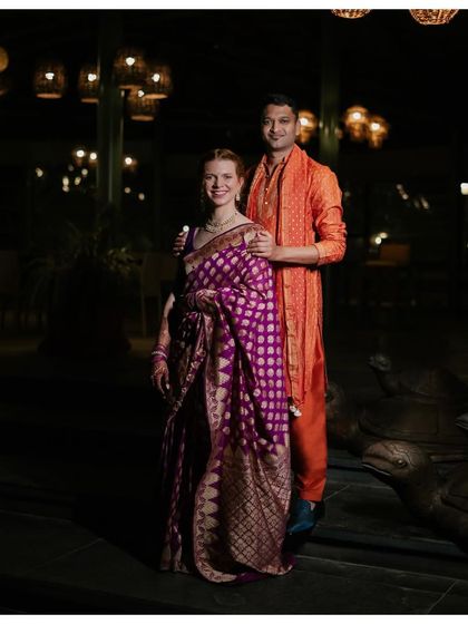 A full-length shot of the couple. The groom's custom orange Patiala and Kurta were designed to echo his father's attire in the original portrait, creating a beautiful contrast with the purple saree.
