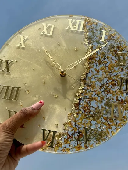 A finished varmala preservation clock, held up to the sky to show the beautiful details of the preserved flowers and gold foil.