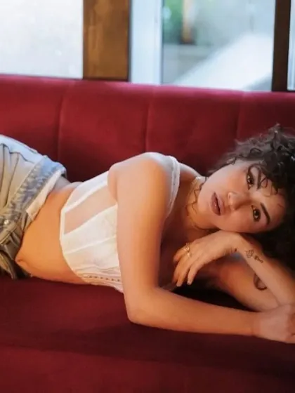 A relaxed, reclining pose that still feels high-fashion. The soft, diffused lighting highlights the texture of her curly hair and the natural-looking finish of the makeup.
