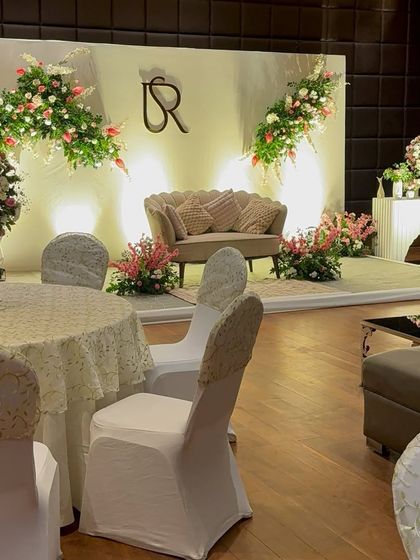 A full view of an elegant Roka stage setup. The design includes a clean white backdrop, asymmetrical floral arrangements, a custom couple's monogram, and a comfortable lounge sofa for the couple.