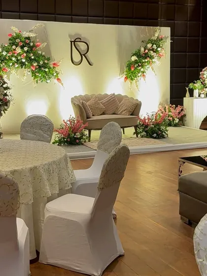A full view of an elegant Roka stage setup. The design includes a clean white backdrop, asymmetrical floral arrangements, a custom couple's monogram, and a comfortable lounge sofa for the couple.