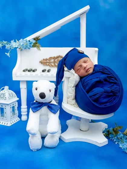 A newborn boy sleeps peacefully on a miniature piano chair, surrounded by a dreamy blue-themed setup with a polar bear prop.