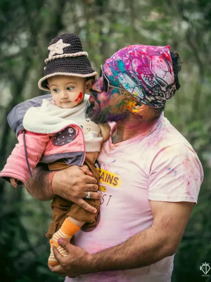A side profile capturing the baby's curious expression as he looks at his father, whose face is covered in vibrant Holi colors. This image tells a story of a shared first experience.