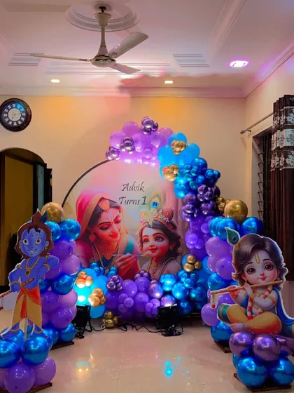 A divine Krishna-themed first birthday decoration. This setup features a circular backdrop with an image of Yashoda and Krishna, flanked by character cutouts and a rich blue and purple balloon garland.