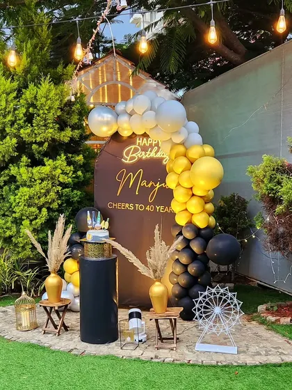 A sophisticated 40th birthday celebration in the garden. The setup includes a personalized backdrop, a black, gold, and silver balloon garland, and elegant decor pieces for a memorable milestone party.
