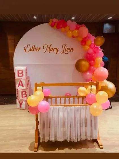 A modern and chic naming ceremony for "Esther Mary Livin". The clean white circular backdrop is accented with a colorful balloon garland, with a traditional wooden cradle at the forefront.