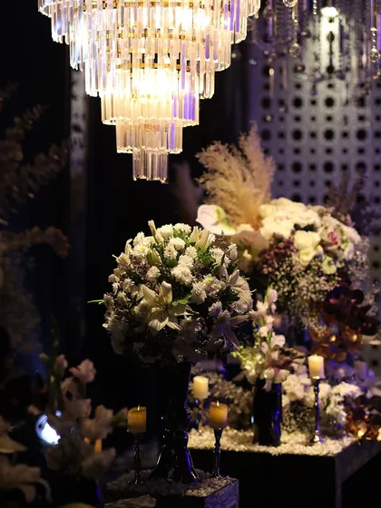 A close-up of a floral arrangement under a crystal chandelier. The mix of white lilies, pampas grass, and other blooms creates a rich, textured look.