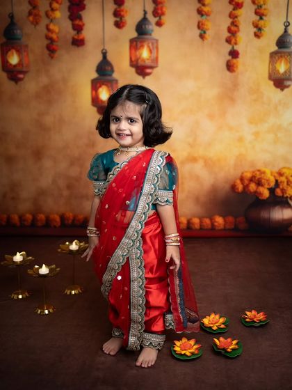 A beautiful portrait of a little girl in a traditional red Maharashtrian saree for her Diwali photoshoot. The warm lighting from the hanging lanterns creates a magical glow.