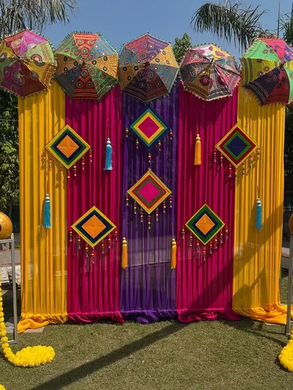 A colorful outdoor Mehendi or Haldi photo booth with a pink and yellow backdrop, decorated with Rajasthani umbrellas and marigold garlands.
