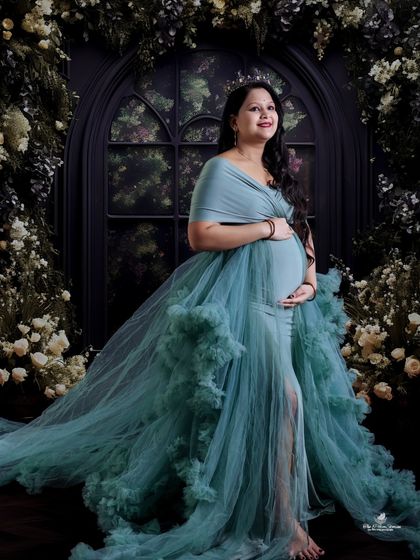 A regal solo portrait. The mom-to-be wears a small crown, standing confidently in a beautiful mint green gown against a dark, enchanting floral backdrop.