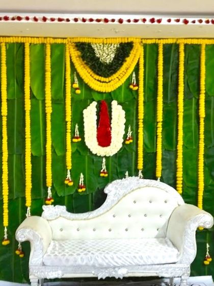 A traditional backdrop for a Half Saree ceremony. The decor features a wall of banana leaves, marigold garlands, and a floral 'Naamam' symbol.