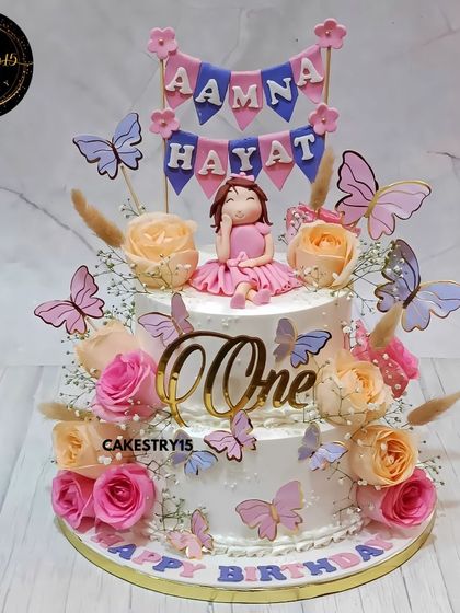 A stunning two-tier chocolate cake for twins' first birthday. This doll-themed cake is elegantly decorated with fresh roses, paper butterflies, and a custom name banner for "Aamna & Hayat".