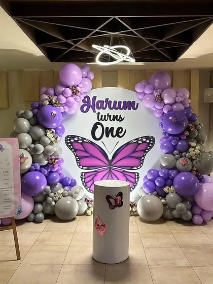 A beautiful butterfly themed first birthday party, with a purple and grey balloon arch framing a large circular backdrop with a monarch butterfly graphic.
