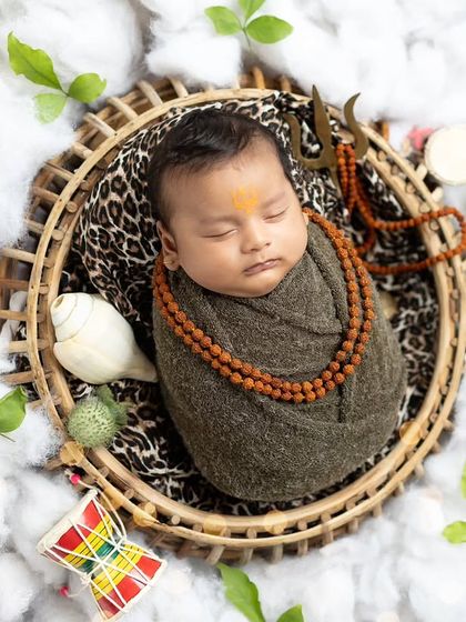 This top-down view shows the complete setup, with the baby nestled in a basket on a bed of clouds, surrounded by belpatra leaves, a shankha, and other divine elements.