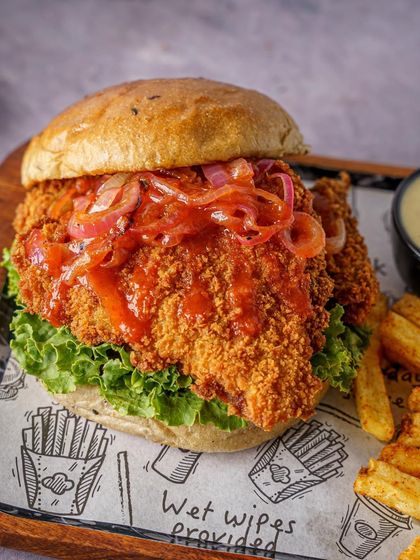 A crispy chicken burger with pickled onions. The focus is on the crunchy texture of the fried chicken patty. The simple styling on a wooden board keeps the attention on the hero product.
