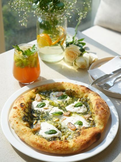 A beautiful shot of our Basil Pesto and Burrata pizza, enjoyed in the sun with a refreshing drink. This is one of our most popular and photogenic dishes.
