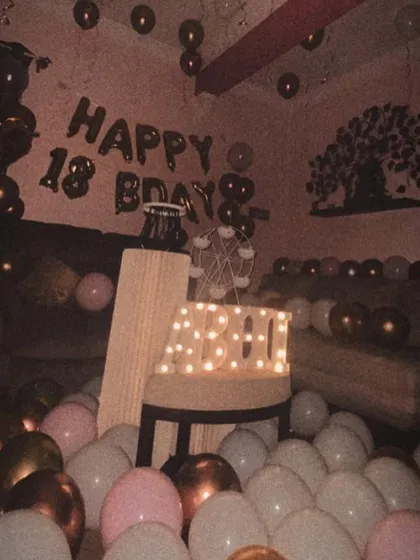 A more muted, vintage-style photo of the 18th birthday room setup, highlighting the warm glow of the marquee letters and the sheer volume of balloons.