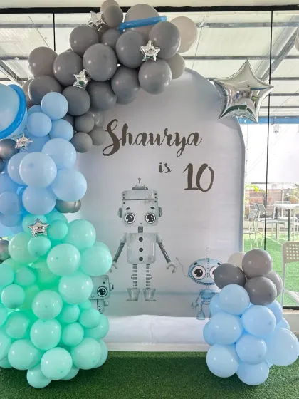 A cool robot-themed 10th birthday party. The setup features a custom backdrop with robot graphics and a balloon garland in shades of mint green, blue, and grey.