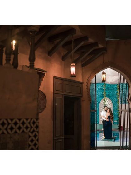 A beautifully layered shot looking through a doorway into an intimate moment by a pool. The warm lanterns and intricate architecture create a rich, moody, and romantic scene.