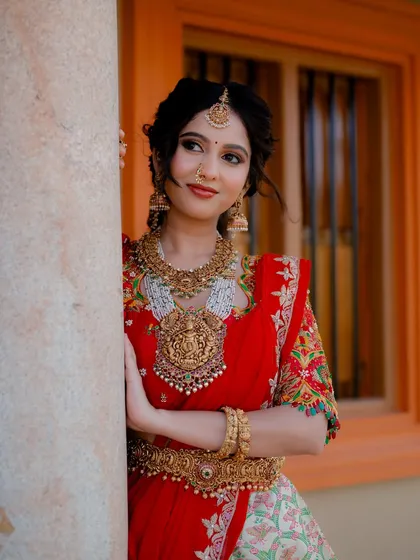 This portrait of actress Niveditha Gowda showcases a stunning red half saree and temple jewelry. The composition highlights the collaboration between the designer, makeup artist, and jeweler.