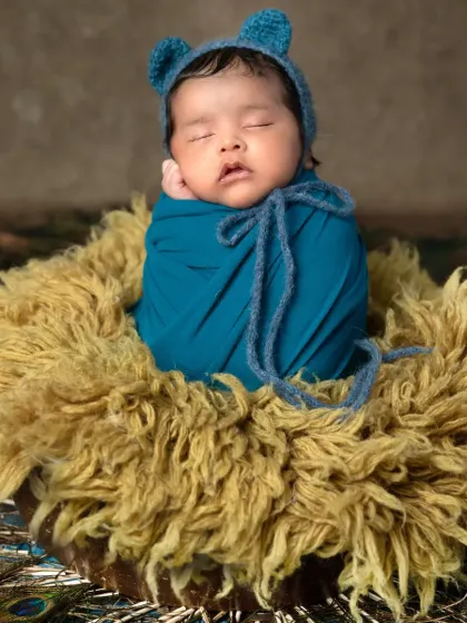 A close-up of a newborn in a knitted blue bonnet, nestled in a fluffy basket. This shot highlights the textures and colors of the Krishna theme, from the soft wool to the vibrant peacock feathers.