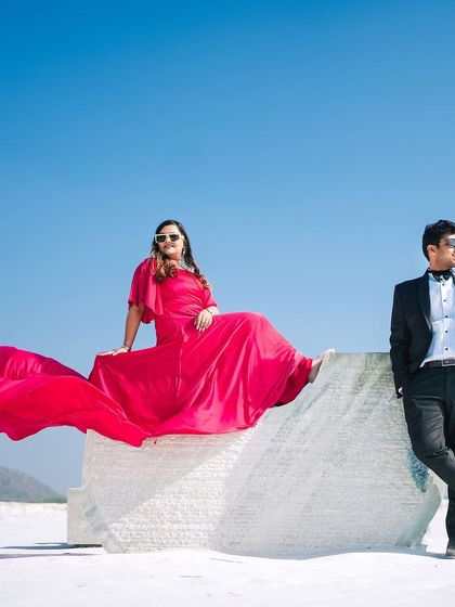 A cool, editorial-style pose against a bright blue sky and white ground. The flowing red dress adds a dynamic element to this modern and stylish pre-wedding shot.