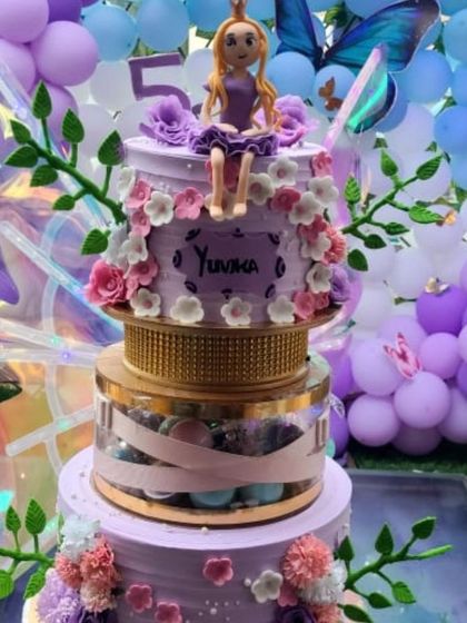 A close-up of a beautiful purple butterfly wing cake, featuring a princess topper and delicate floral decorations. The clear tier is filled with macarons, adding another layer of detail.