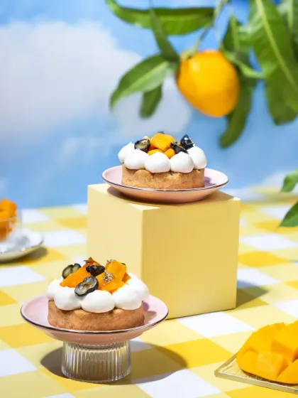 Two beautiful mango tarts with whipped cream and fresh fruit, styled with geometric blocks. The playful and artistic setup with a summer sky backdrop is perfect for a creative dessert menu.