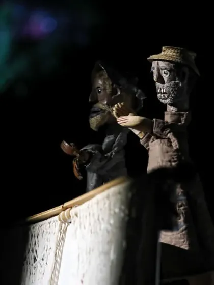 Two skeletal-looking puppets interact in a darkly lit scene from 'Mamulengo de la Mancha', showcasing the show's witty and satirical tone.