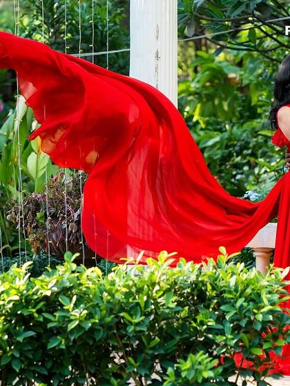 A couple shares an intimate moment in a garden, her flowing red gown adding a vibrant splash of color and drama to the natural surroundings.