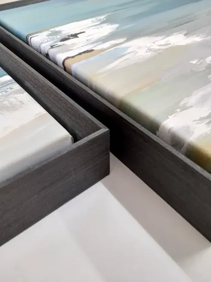 A detail shot of two coastline paintings in floater frames. The dark wood-look finish provides a strong border that contains the energy of the abstract seascapes.