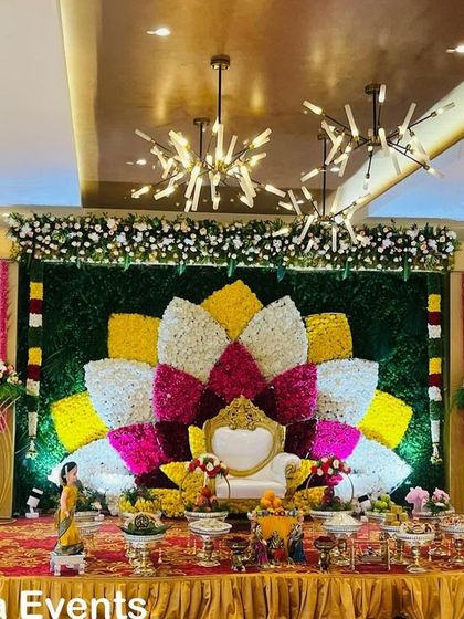 A wide view of a grand hall decorated for a Seemantham ceremony. The stage features a large, multi-colored floral lotus design against a green backdrop, creating a spectacular focal point.