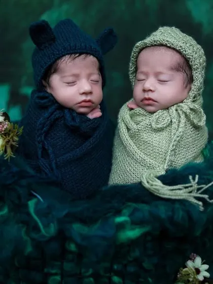 These newborn twins are styled in adorable knit bonnets, one in navy blue and one in sage green, nestled together in a dark, moody, forest-themed setup.